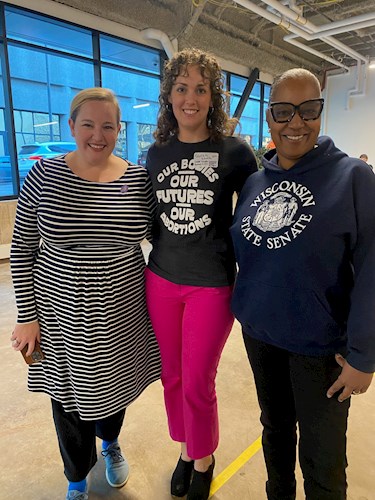 Three women stand together. Senator Kelda Roys is wearing a black and white striped dress, Supervisor Sarah Smith is wearing a black shirt with white print and pink pants, and Senator Lena Taylor is wearing a blue sweatshirt and black pants. 